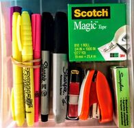 A neatly organized drawer of office supplies, including highlighters, mini stapler and staples, tape, permanent markers, and adhesive tabs.